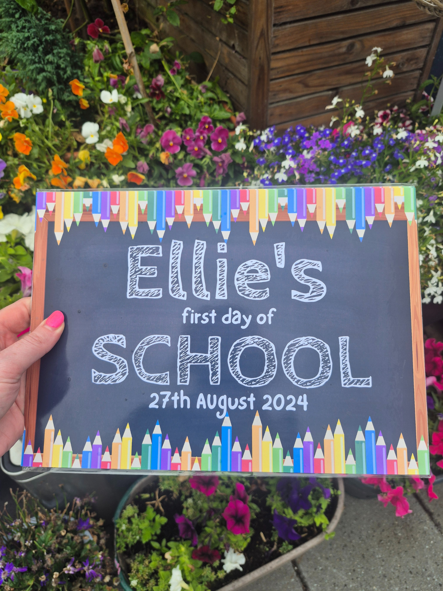 Decorative sign for 'Ellie's first day of school' with colorful crayon border, held in front of flowers.