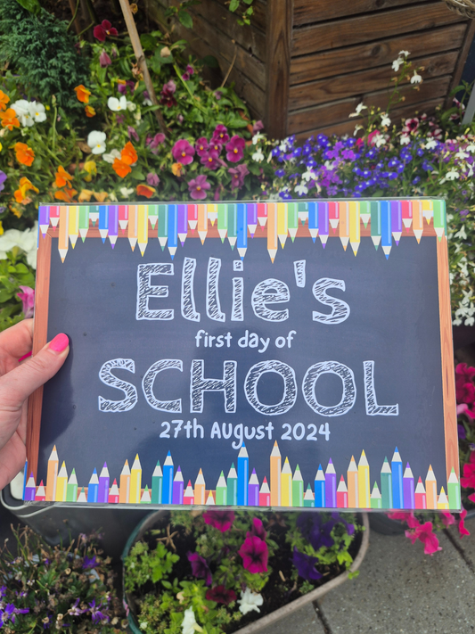 Decorative sign for 'Ellie's first day of school' with colorful crayon border, held in front of flowers.